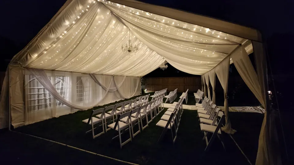 A white event tent dressed in ceiling draping and warm twinkle lights at night