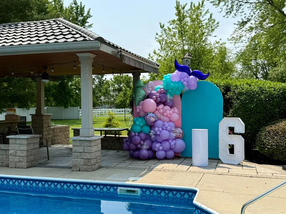 A turquoise backdrop with lavender, pink and aqua balloons and a mermaid tail beside a pool