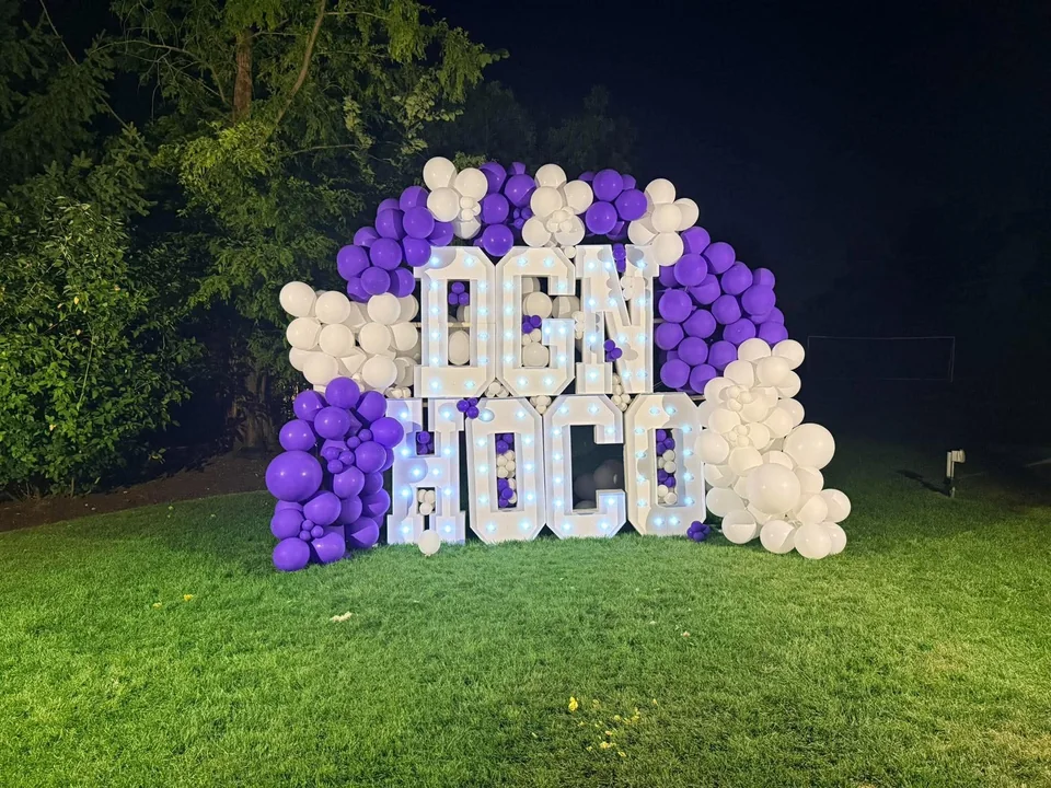 Large illuminated school homecoming letters surrounded by purple and white balloons on a lawn at night